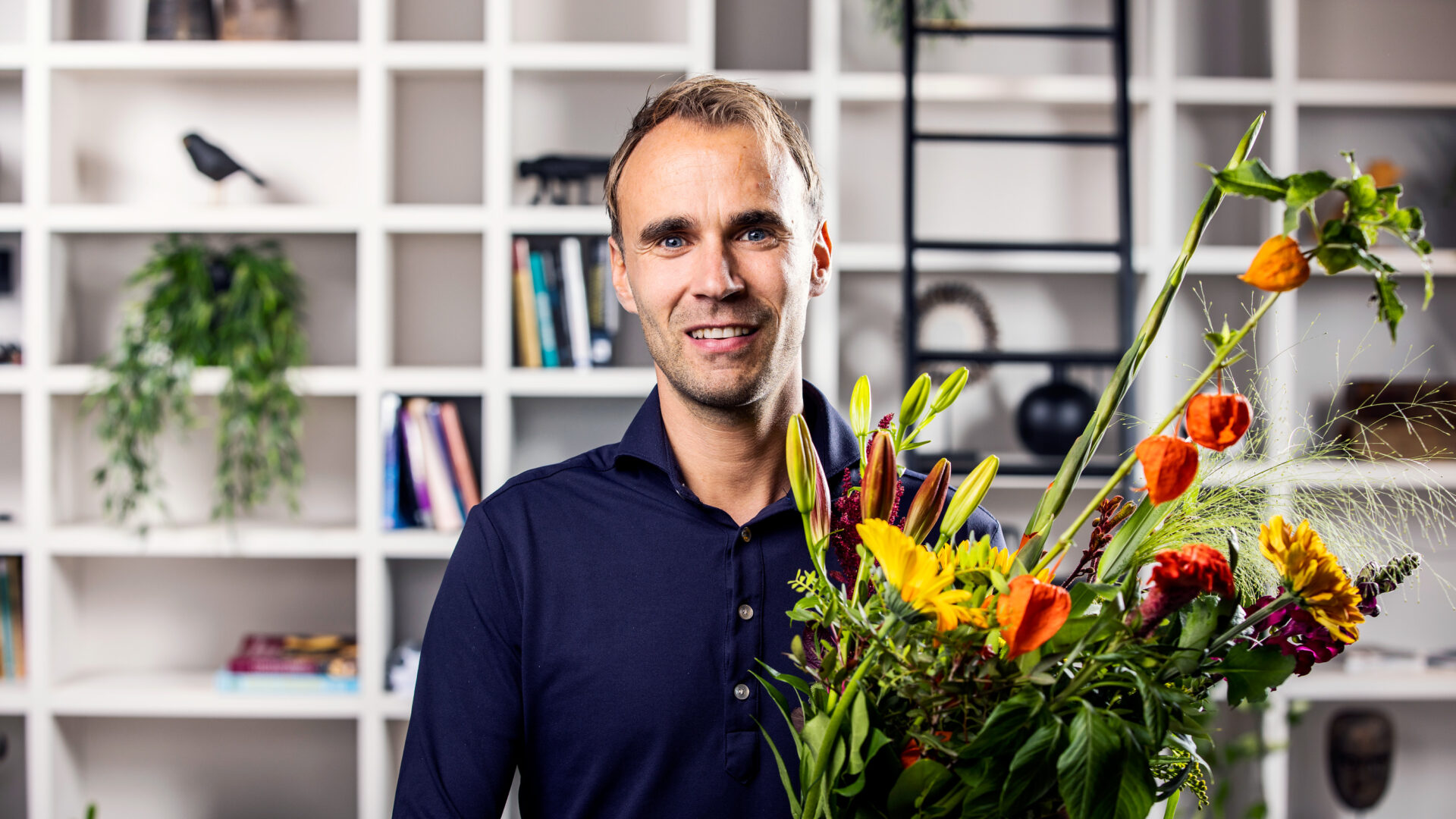 Man die glimlachend een kleurrijk boeket bloemen vasthoudt, staand voor een wandkast met planten en decoratie.